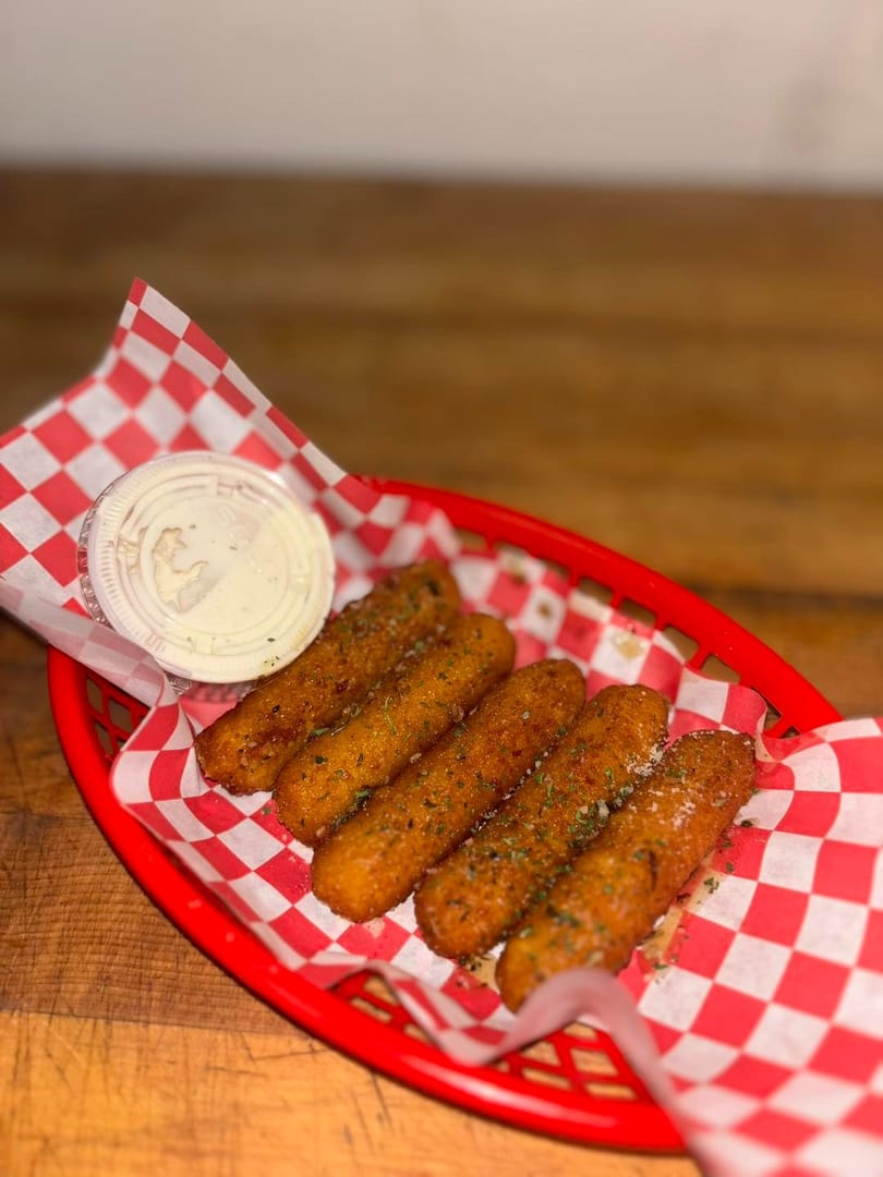 Five golden-brown mozzarella sticks with herbs and a side of ranch in a red basket with checkered paper.