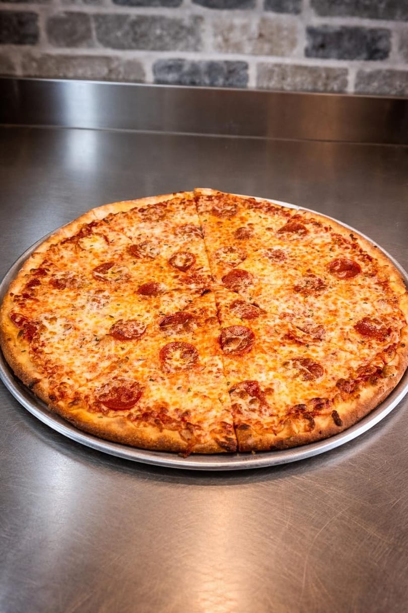 A whole pepperoni pizza, sliced into eight pieces, on a metal tray with a brick wall background.