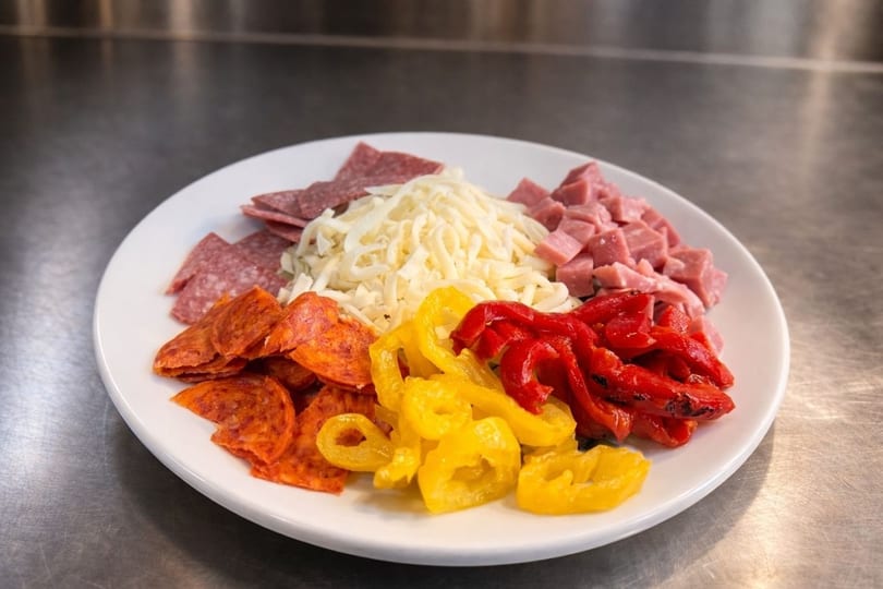 A white plate with pizza toppings: pepperoni, salami, ham, shredded cheese, yellow and red peppers.