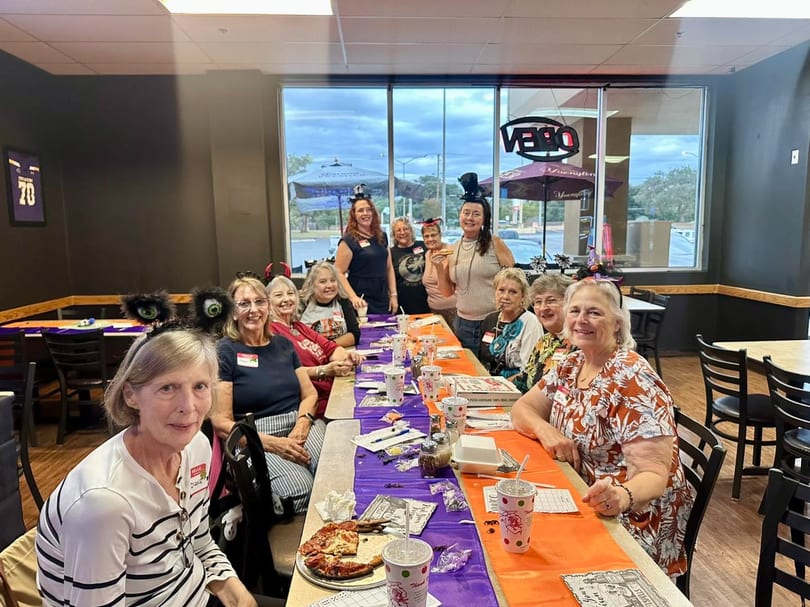 A group of women in Halloween headbands and costumes sit at a long table with pizza and drinks.