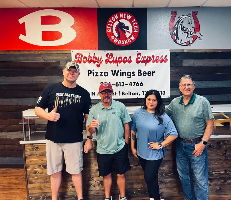 Four people stand in front of a Bobby Lupos Express sign, with a wooden wall behind them.