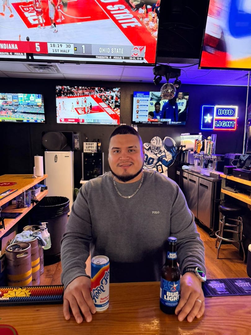 Man sitting at a bar with drinks, surrounded by multiple TVs showing sports games.