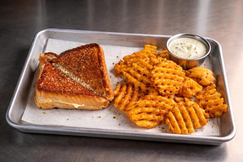 Grilled cheese sandwich with waffle fries and a side of dipping sauce on a metal tray.