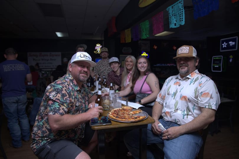 Group of people enjoying pizza and drinks at a dimly lit restaurant with colorful decorations.