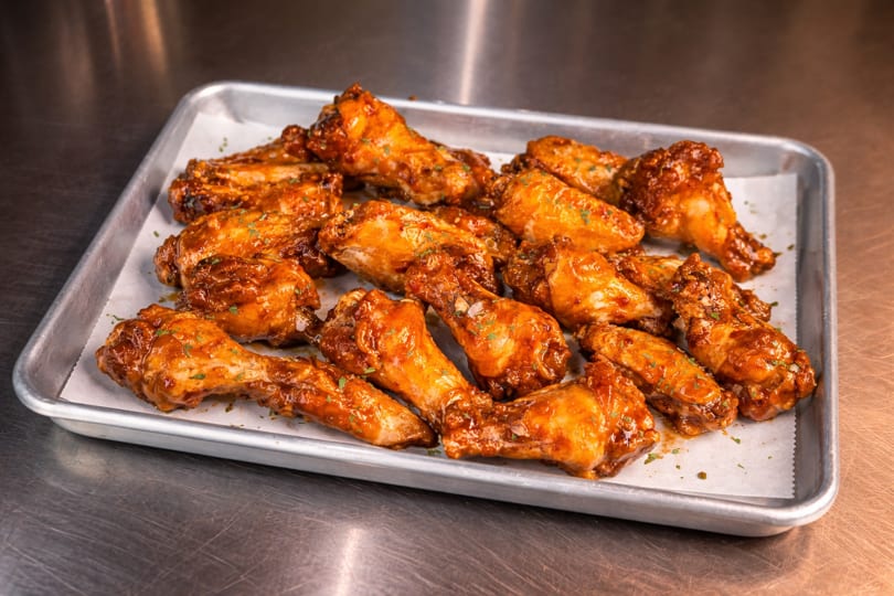 A metal tray filled with numerous glossy, sauced chicken wings on white parchment paper.
